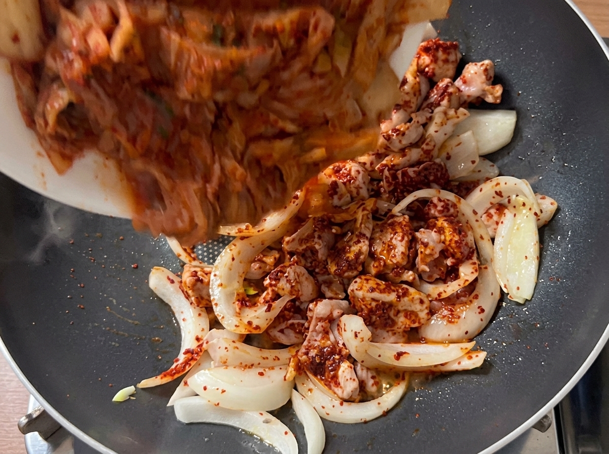 炒めた豚肉と玉ねぎに、一口大に切ったキムチを加えている様子。