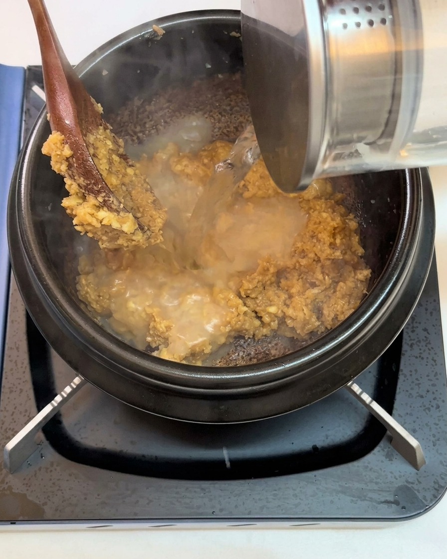 炒めた味噌とにんにくが入った鍋に水を注いでいる様子。