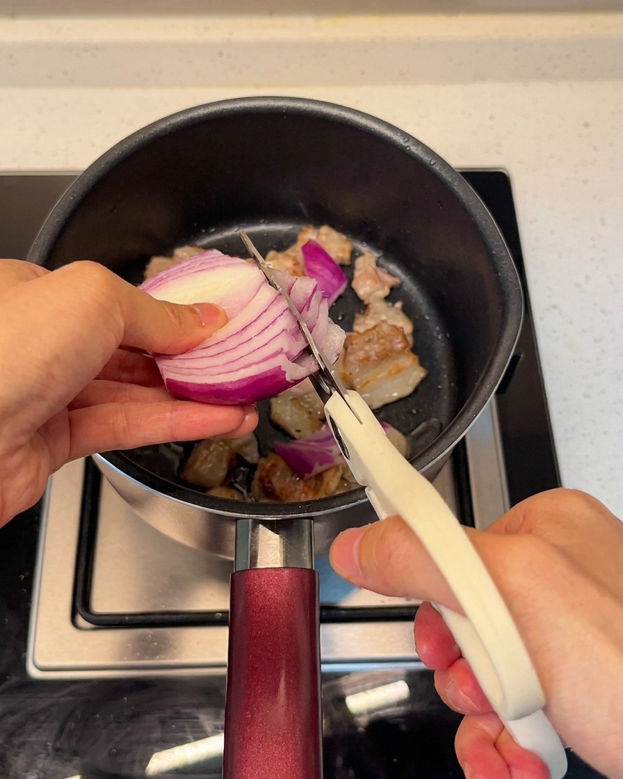 焼いた豚肉の入った鍋に、キッチンバサミで玉ねぎを直接切り入れている様子。