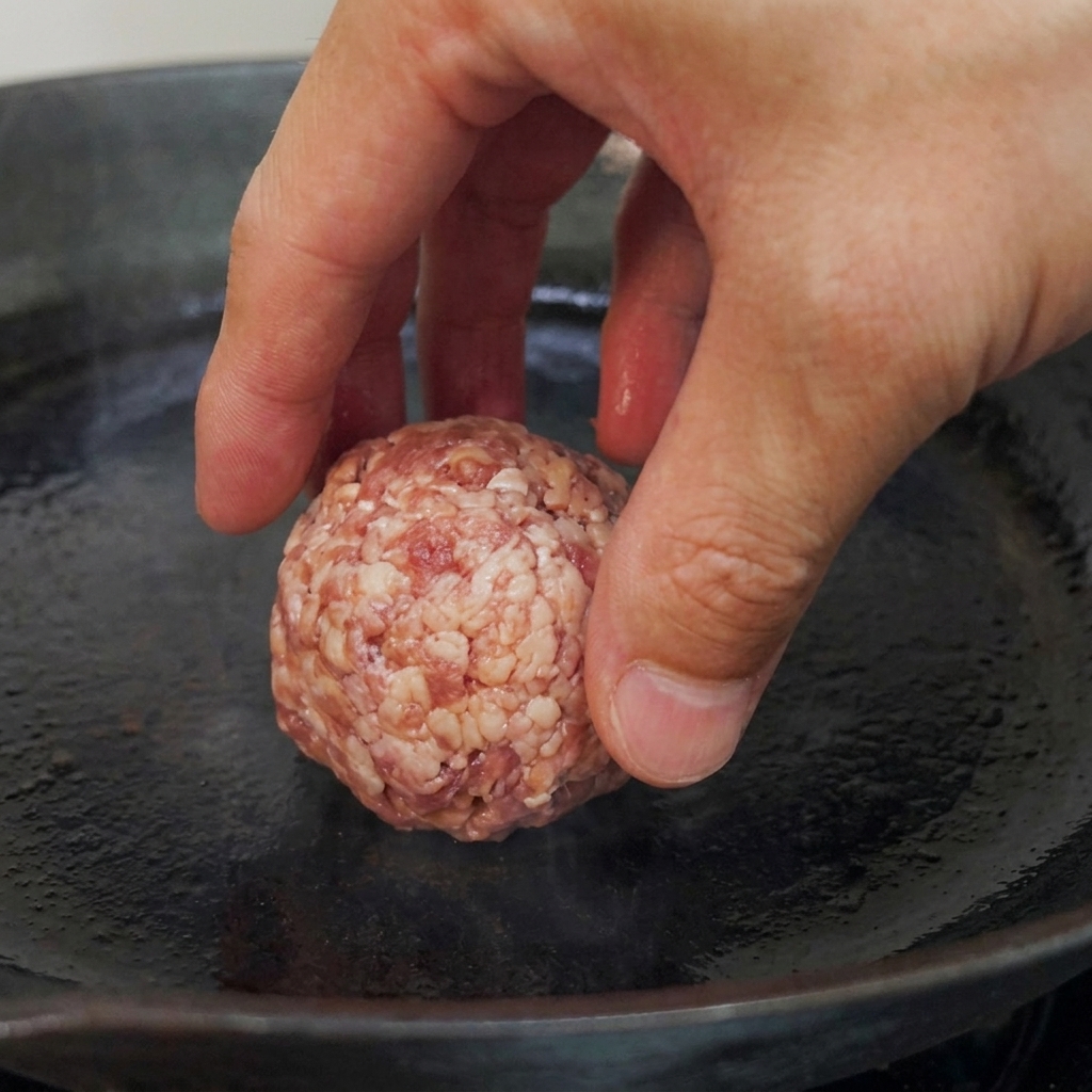 熱した平らなフライパンに生肉のボールを置いている様子。
