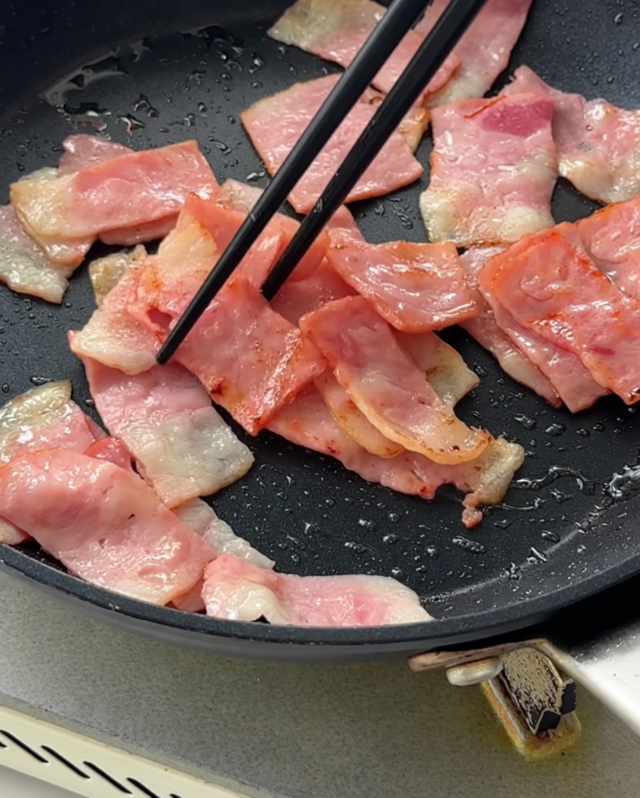 焼けたベーコンが入ったフライパンに刻んだ紫玉ねぎを加えて混ぜている様子。