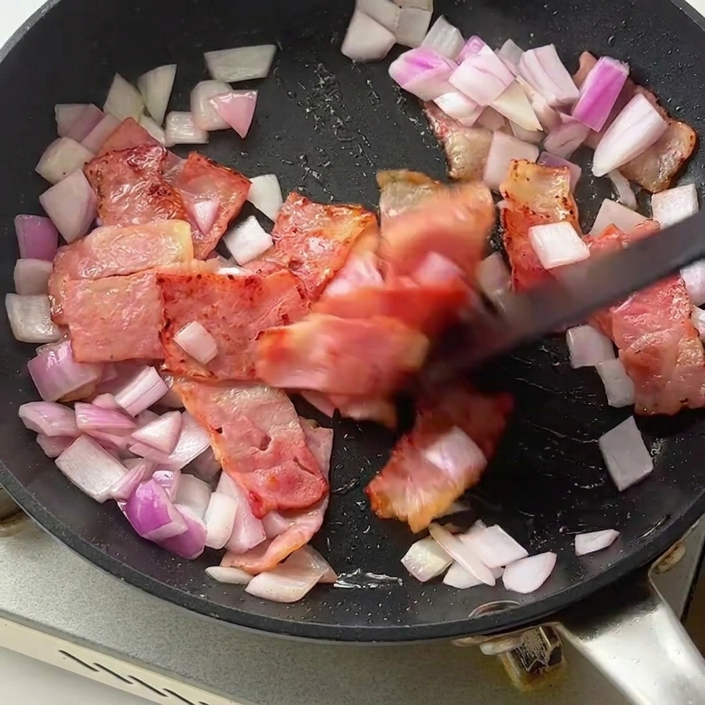 カリカリのベーコンと玉ねぎが入ったフライパンに、茹でたパスタとアスパラガスを加えている様子。