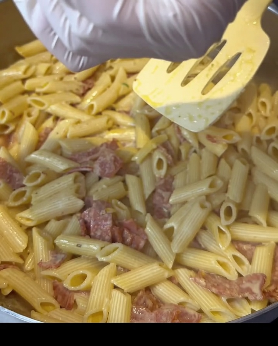 クリーミーなベーコンパスタが入ったフライパンの上から、手袋をした手で調味料を振りかけている様子。