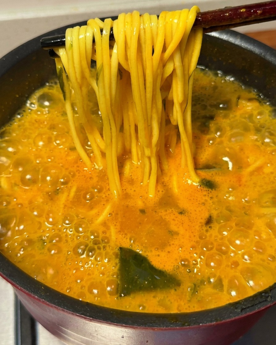 グツグツ煮立つスパイシーなスープから、茹で上がった黄色い麺を箸で持ち上げている様子。