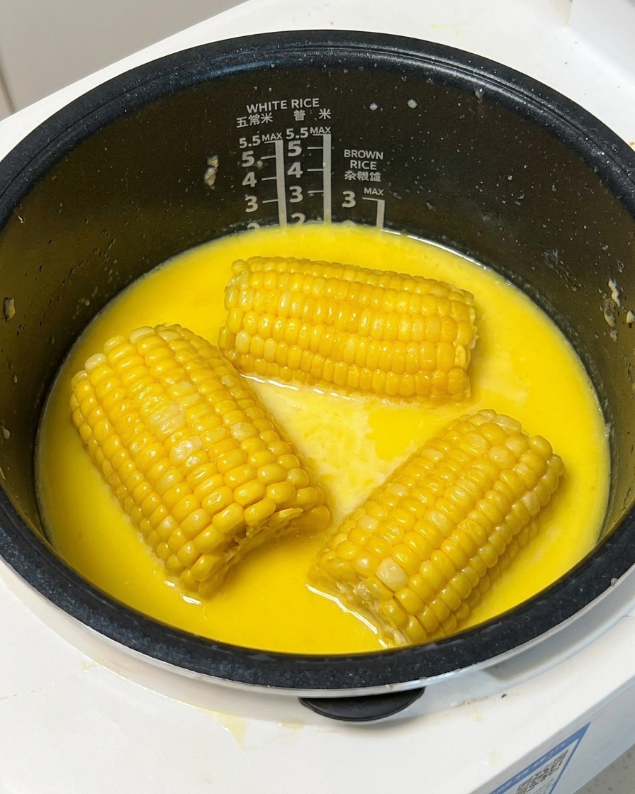 炊飯釜の中のミルキーなスープから、調理済みのコーンを木製串で持ち上げている様子。