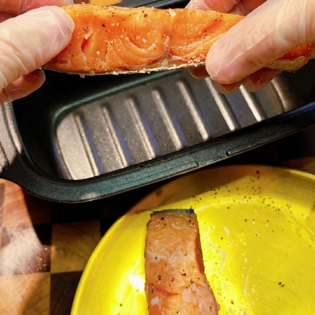 手袋をした手で、下味をつけたサーモンを黒い長方形の電子レンジ用焼き目付けパンに乗せている様子。