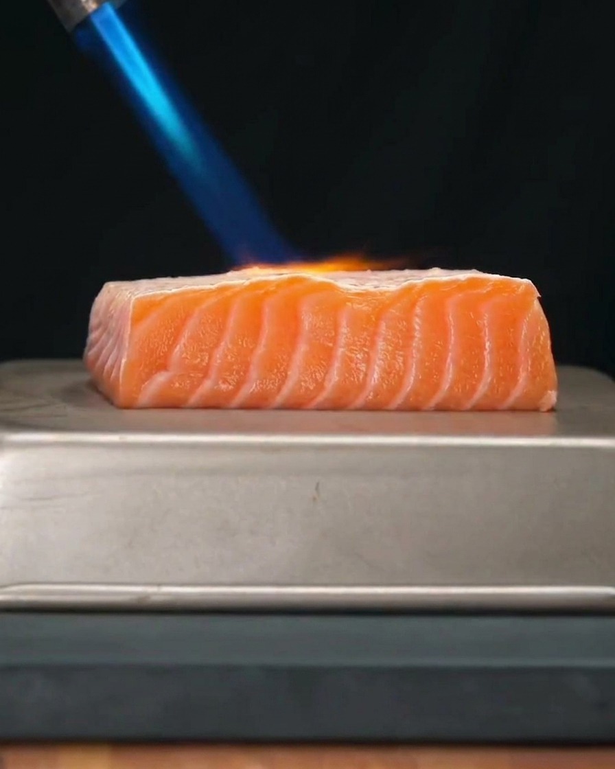 キッチン用バーナーの青い炎で、厚みのある新鮮なサーモンの切り身を炙っている様子。