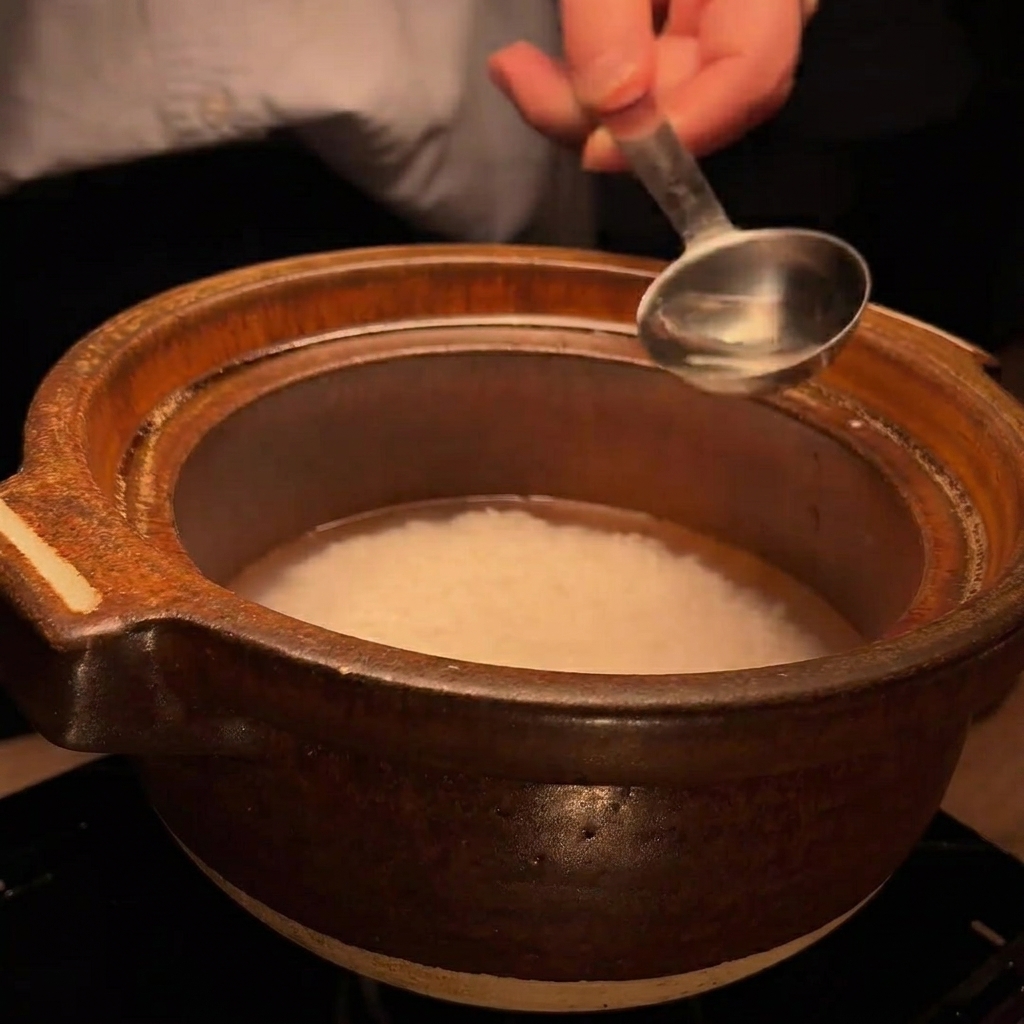 金属のスプーンで酒を土鍋の中の米に加えている様子。