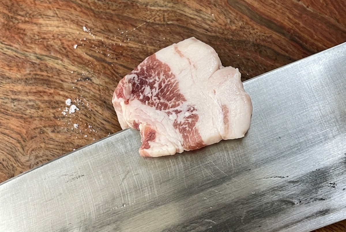 まな板の上で、豚バラ肉の塊を鋭利な包丁で均一な薄切りにしている様子。