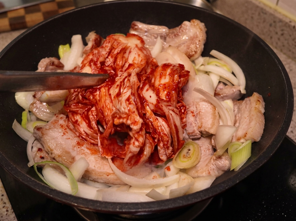 トングを使って、焼いた豚肉と玉ねぎの上に大きなキムチを置く様子。
