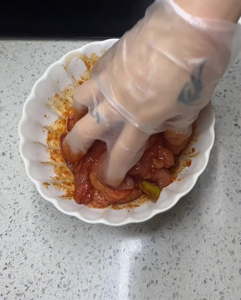 手袋をした手で、ボウルに入った鶏肉に赤いスパイスダレを揉み込んでいる様子。