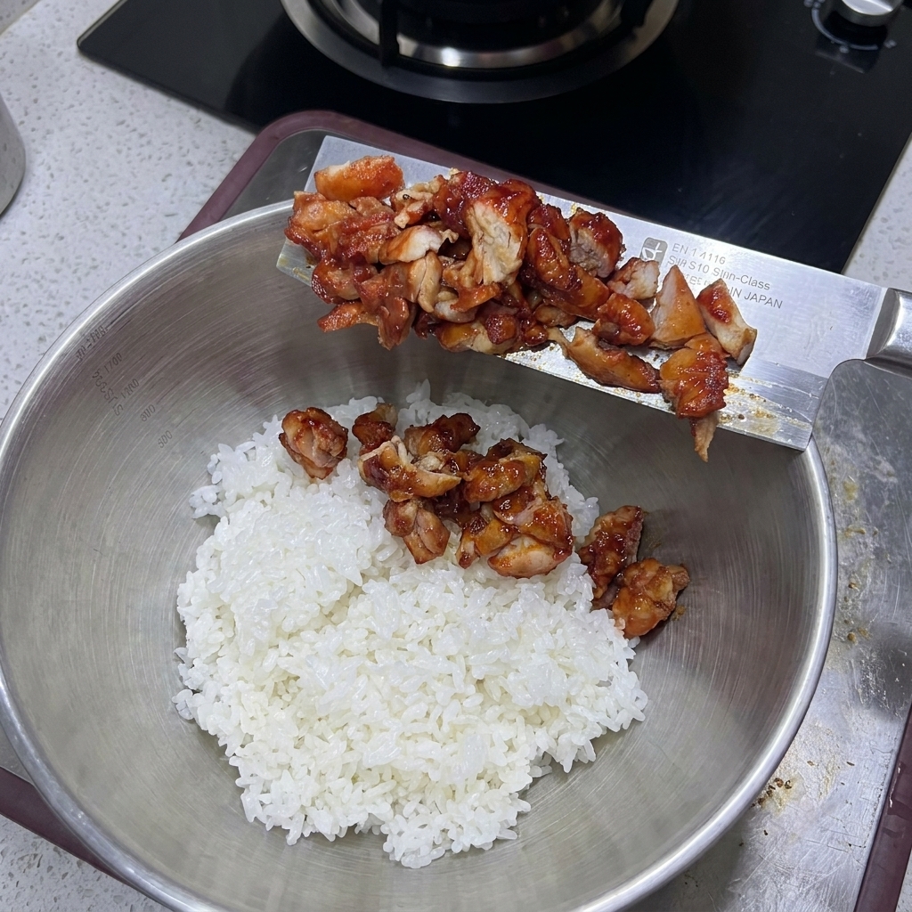炊きたてのご飯が入った大きな金属ボウルに、包丁からカットしたチキンを入れている様子。