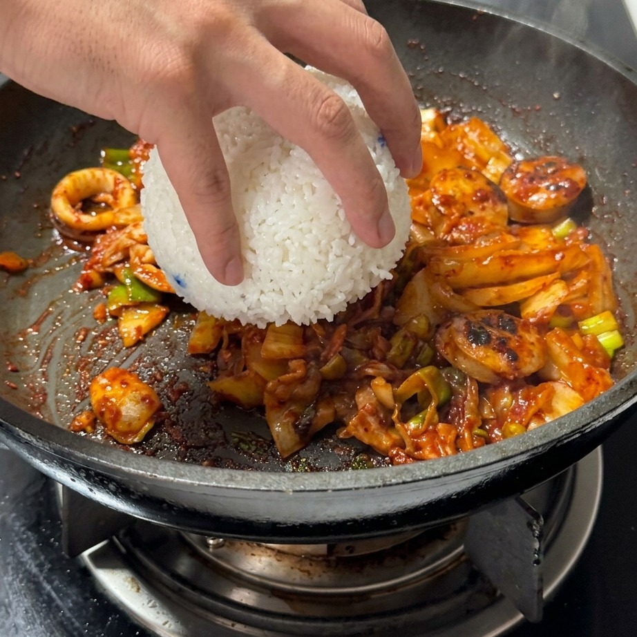 炒めたキムチとお肉が入ったフライパンに、お茶碗一杯の温かい白ご飯を加えている様子。