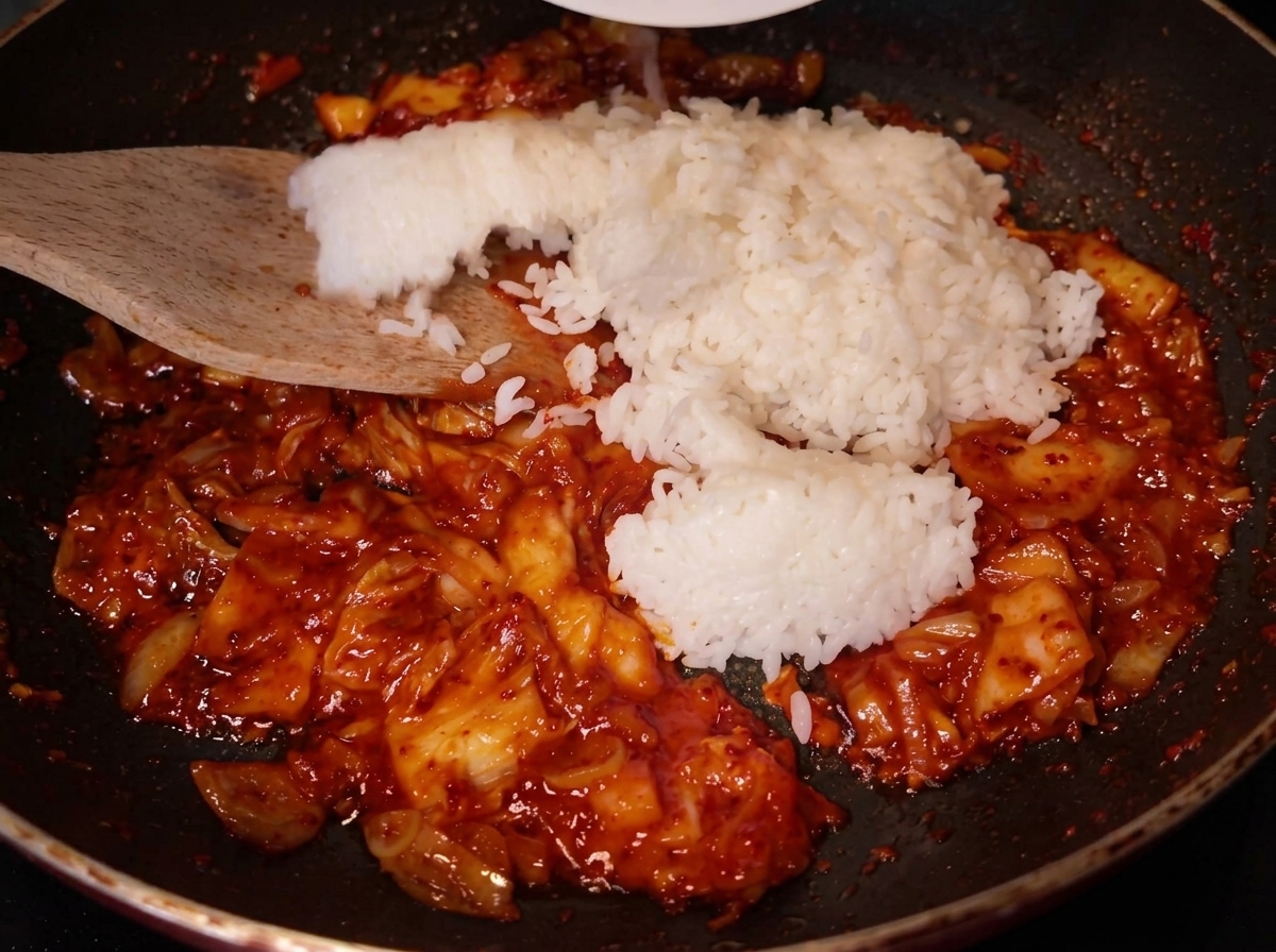 赤いキムチを炒めているフライパンに、木製のヘラでたっぷりの白ご飯を加えている様子。