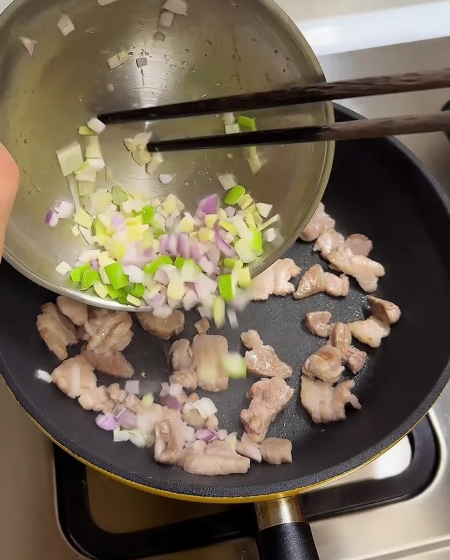 炒めた豚肉の入ったフライパンに、ボウルから青ねぎ、にんにく、玉ねぎを加える様子。