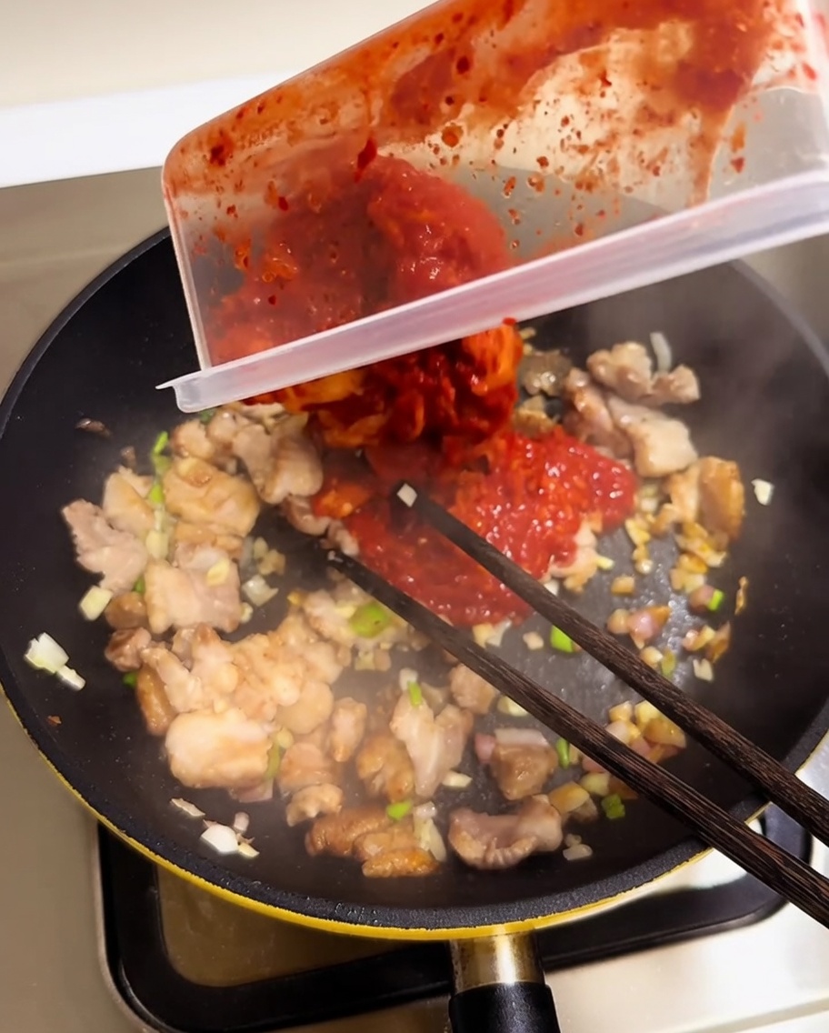 容器から刻んだ赤いキムチを、豚肉と香味野菜の入ったフライパンに加える様子。