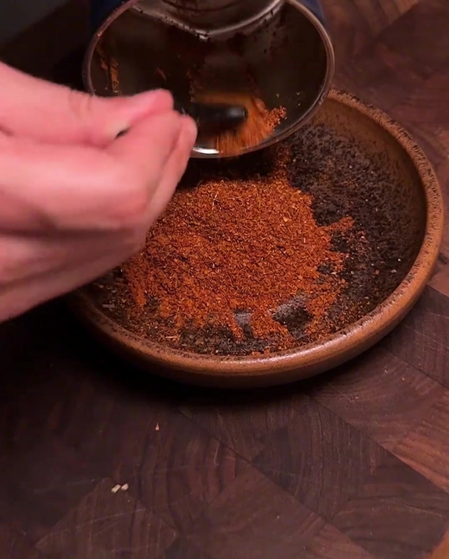 スパイスミルから、挽きたての鮮やかな赤いチリとスパイスの粉末を茶色いボウルに移している様子。