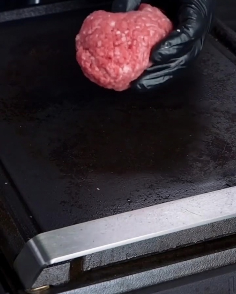 手袋をした手で、生の牛ひき肉を丸めて熱い鉄板の上に置いている様子