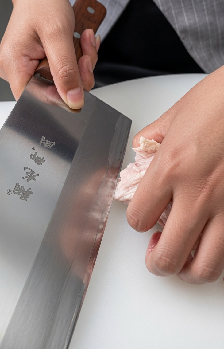 白いまな板の上で、包丁を使って鶏もも肉から骨を切り離している様子。