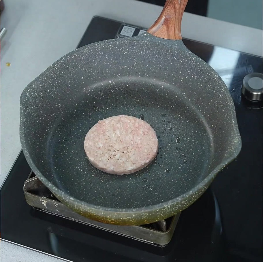 薄く油をひいたフライパンの中で、生のビーフパティがジューシーな音を立てて焼き始めている様子。