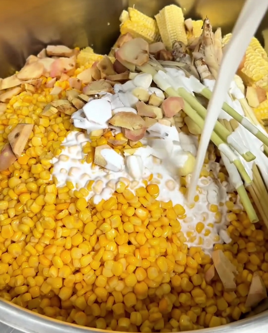 鍋の中のとうもろこしと薬味の上に、白いココナッツミルクととうもろこしの芯を入れている様子。