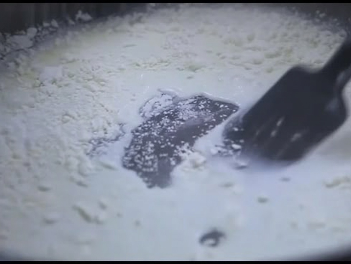 鍋の中の白くとろみのあるミルクと小麦粉の混合物を、黒いシリコンスパチュラでかき混ぜている様子。