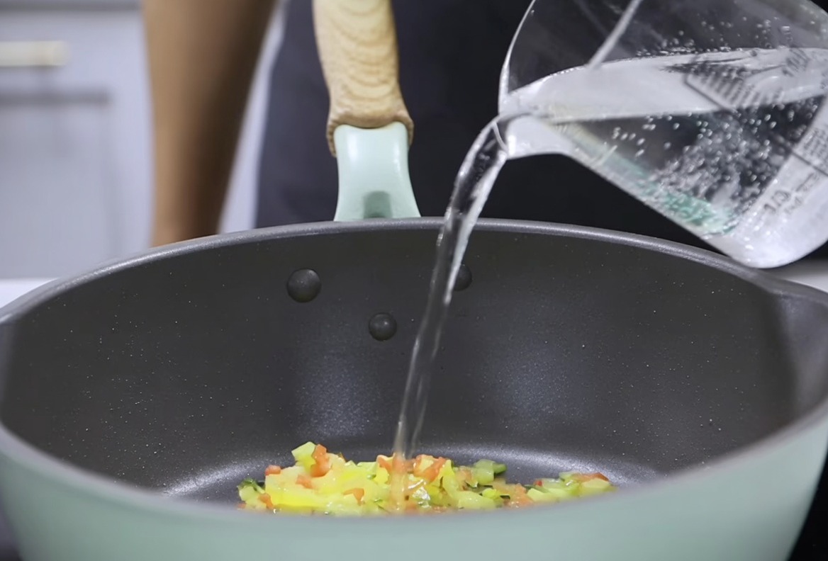 炒めた野菜の入った鍋に計量カップから水を注ぎ、とろみをつけたミルクのルーを加える準備をしている様子。