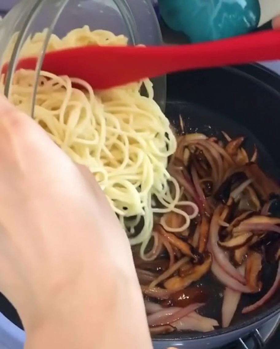 味付けされたきのこと玉ねぎのフライパンに、茹でたスパゲッティをボウルから加えているところ。