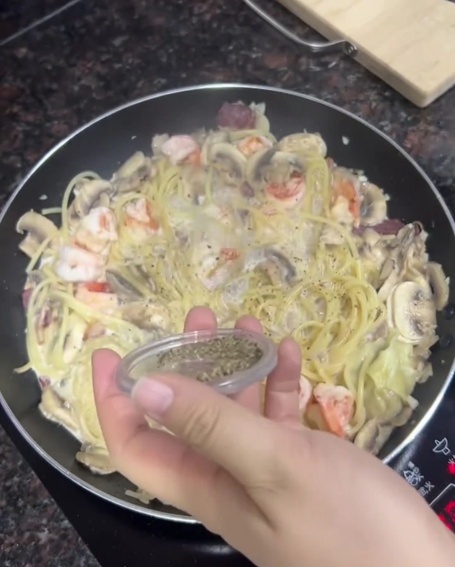 クリーミーなパスタ、エビ、マッシュルームが入ったフライパンに、手で粗挽き黒こしょうを振りかけている様子。