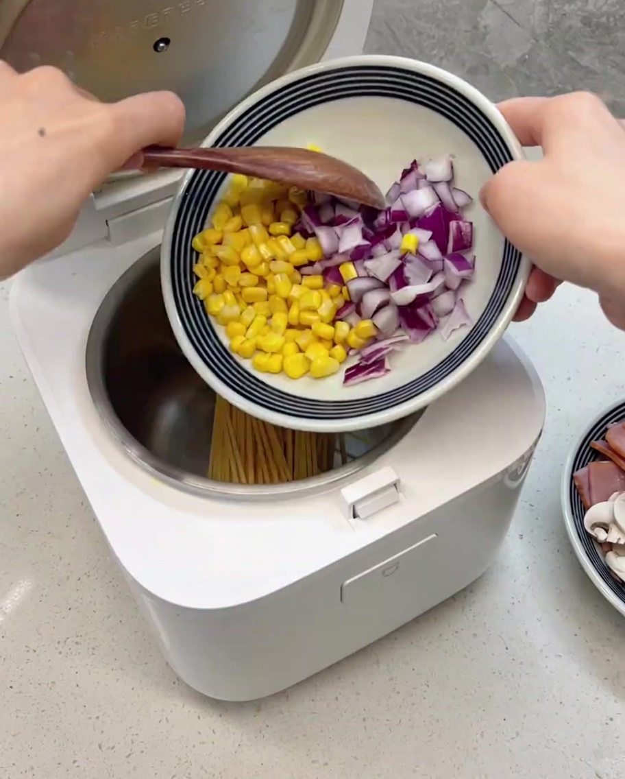 炊飯器のパスタの上にコーン、紫玉ねぎ、ハム、マッシュルームなどの具材を乗せた状態。