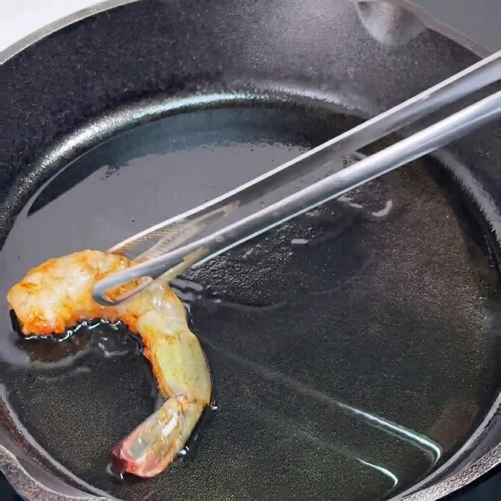 熱したスキレットの上で、トングを使って半分火の通ったエビをひっくり返している様子。