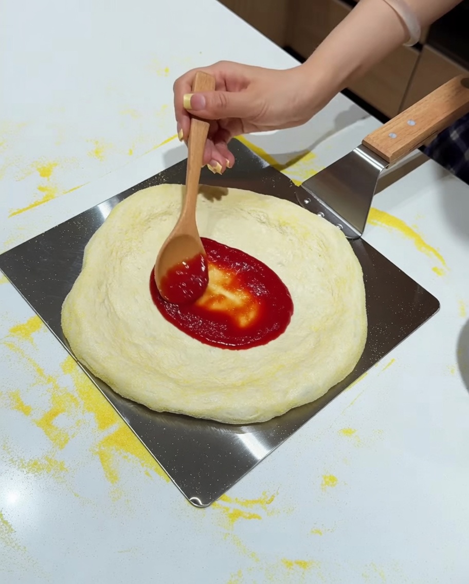 メタルピールの上で、木のスプーンを使ってトマトソースを塗られるピザ生地。