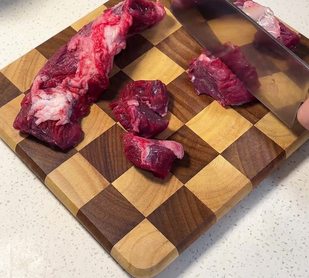 市松模様の木製まな板の上で、牛バラ肉を均等な角切りにしている様子。