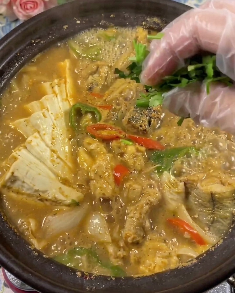 グツグツ煮え滾るタラコと豆腐の土鍋スープに、透明な手袋をした手で新鮮なパクチーをのせているクローズアップ画像。