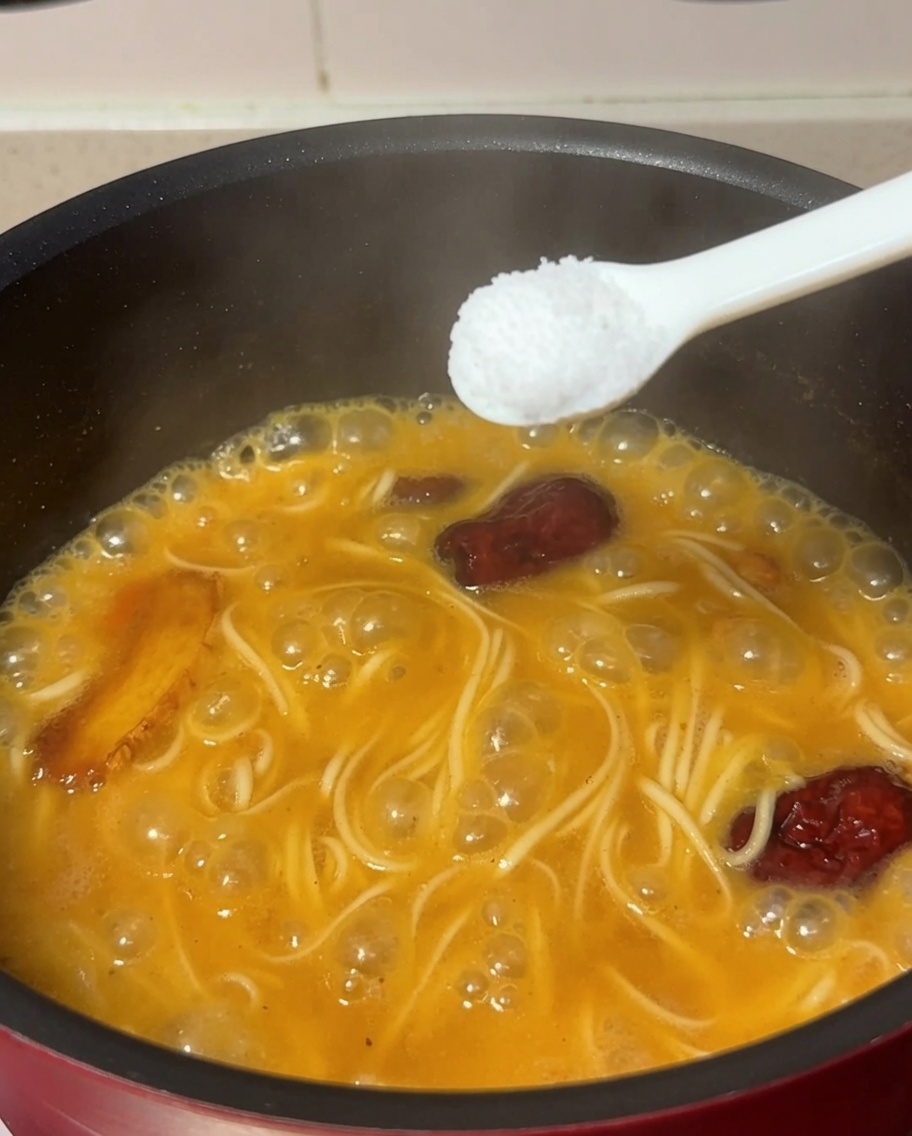 茹でた麺と鶏肉スープの入った鍋に、スプーン一杯の塩を加えている様子。