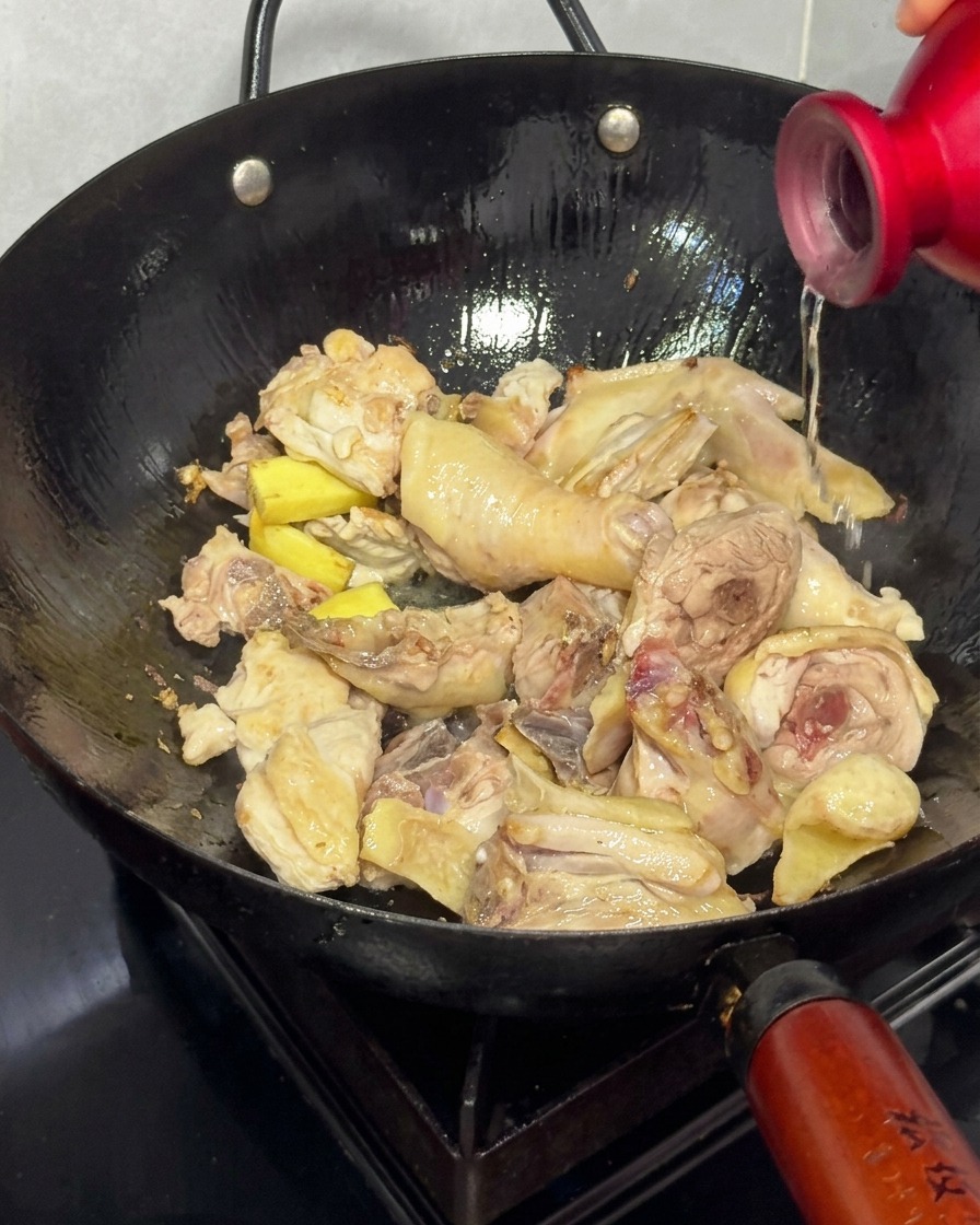 こんがり焼けた鶏肉と生姜が入った中華鍋に、赤いボトルから料理酒を注ぐ様子。