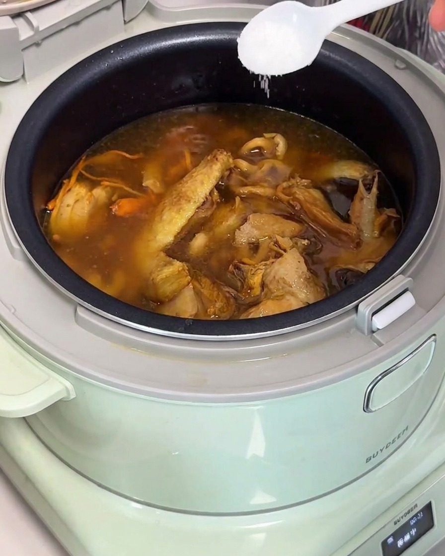 炊飯器の中の完成した黄金色の鶏ときのこスープに、白いスプーンで塩を加える様子。