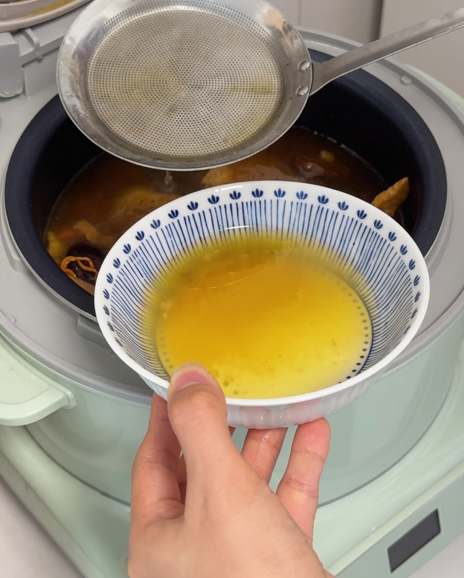 金属製のおたまを使って、炊飯器のスープの表面から黄金色の鶏の脂をすくい取るクローズアップ。