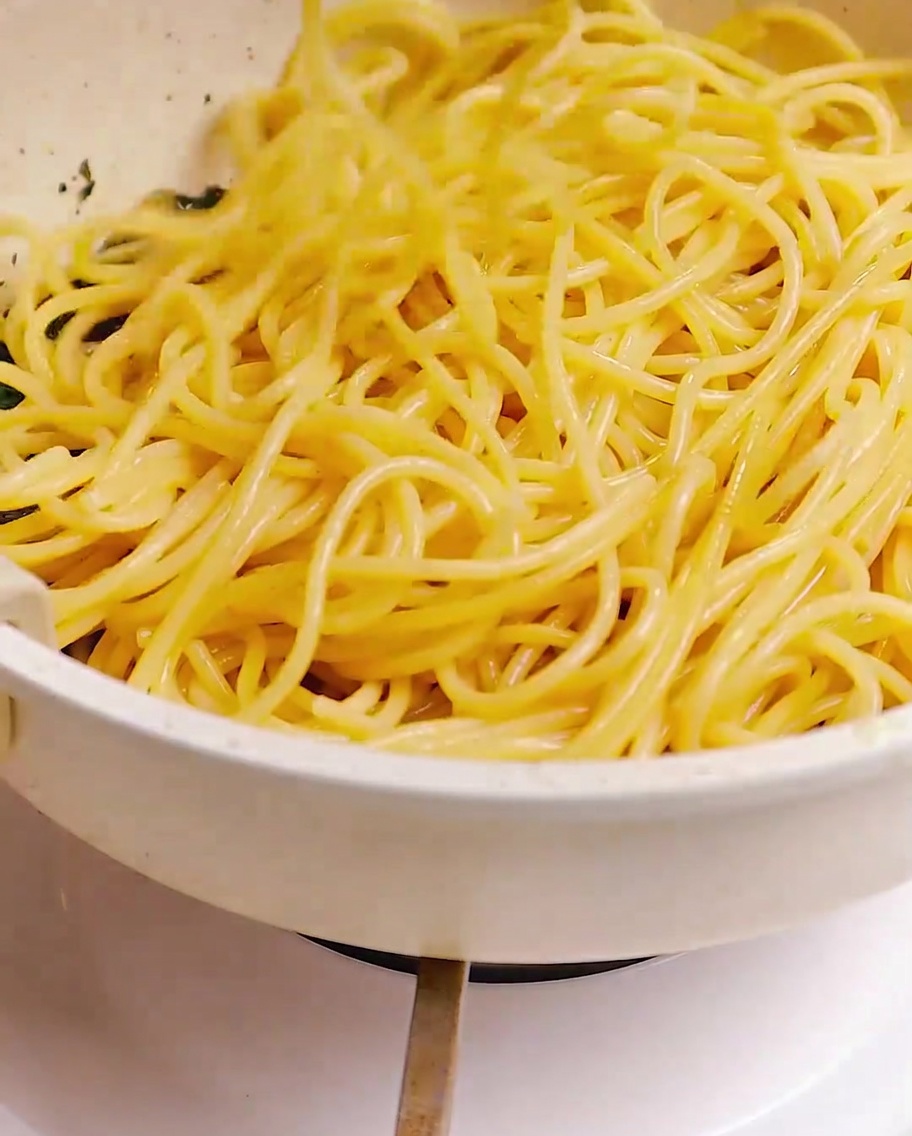 茹で上がったスパゲッティが、フライパンの中でベーコン、ニンニク、ハーブと菜箸で混ぜ合わされている様子。