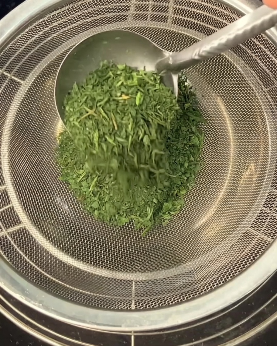 金属製のスプーンを使って、砕いた乾燥セロリの葉を目の細かいザルでボウルにこし入れている様子。