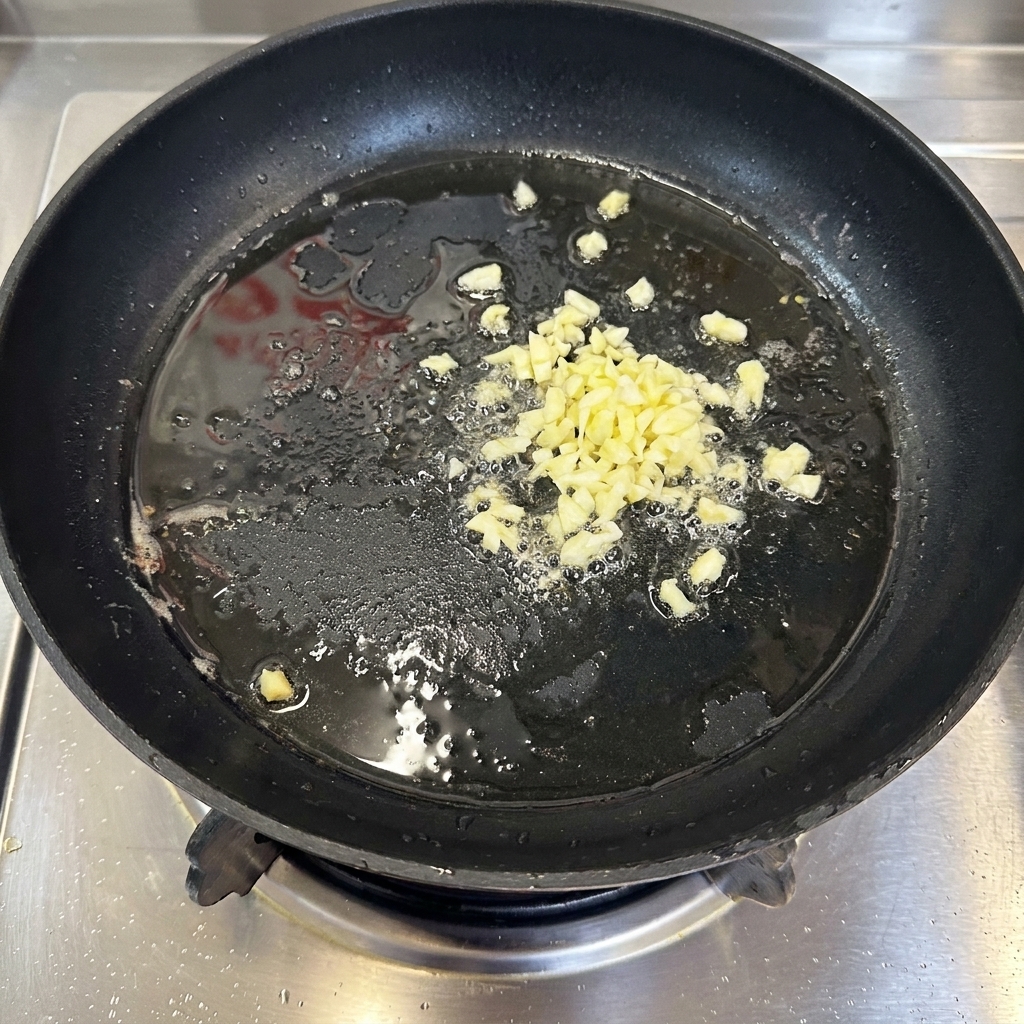 黒いフライパンの中で、薄くひいた熱い油でみじん切りにしたにんにくが軽く泡立って炒められている様子。