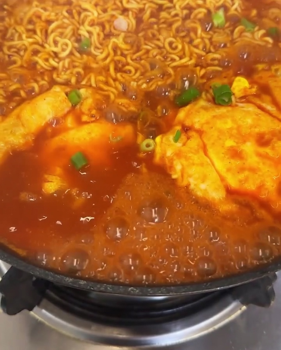 キムチラーメンの鍋に刻んだ青ねぎと鮮やかな赤唐辛子のスライスを散らし、周囲でスープが泡立っているクローズアップ写真。