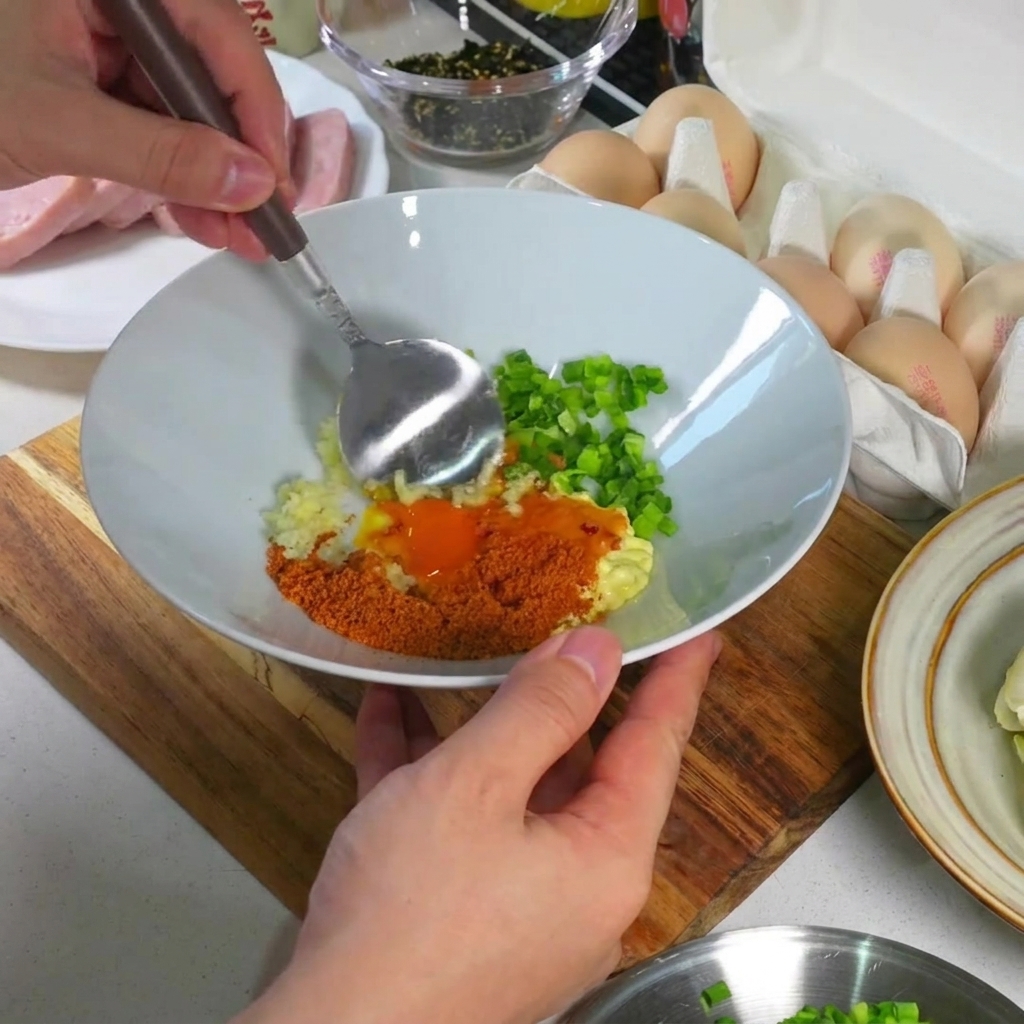 白いボウルの中で、卵黄、ニンニク、ねぎ、調味料をスプーンで滑らかで濃厚なペースト状に混ぜ合わせているクローズアップ写真。