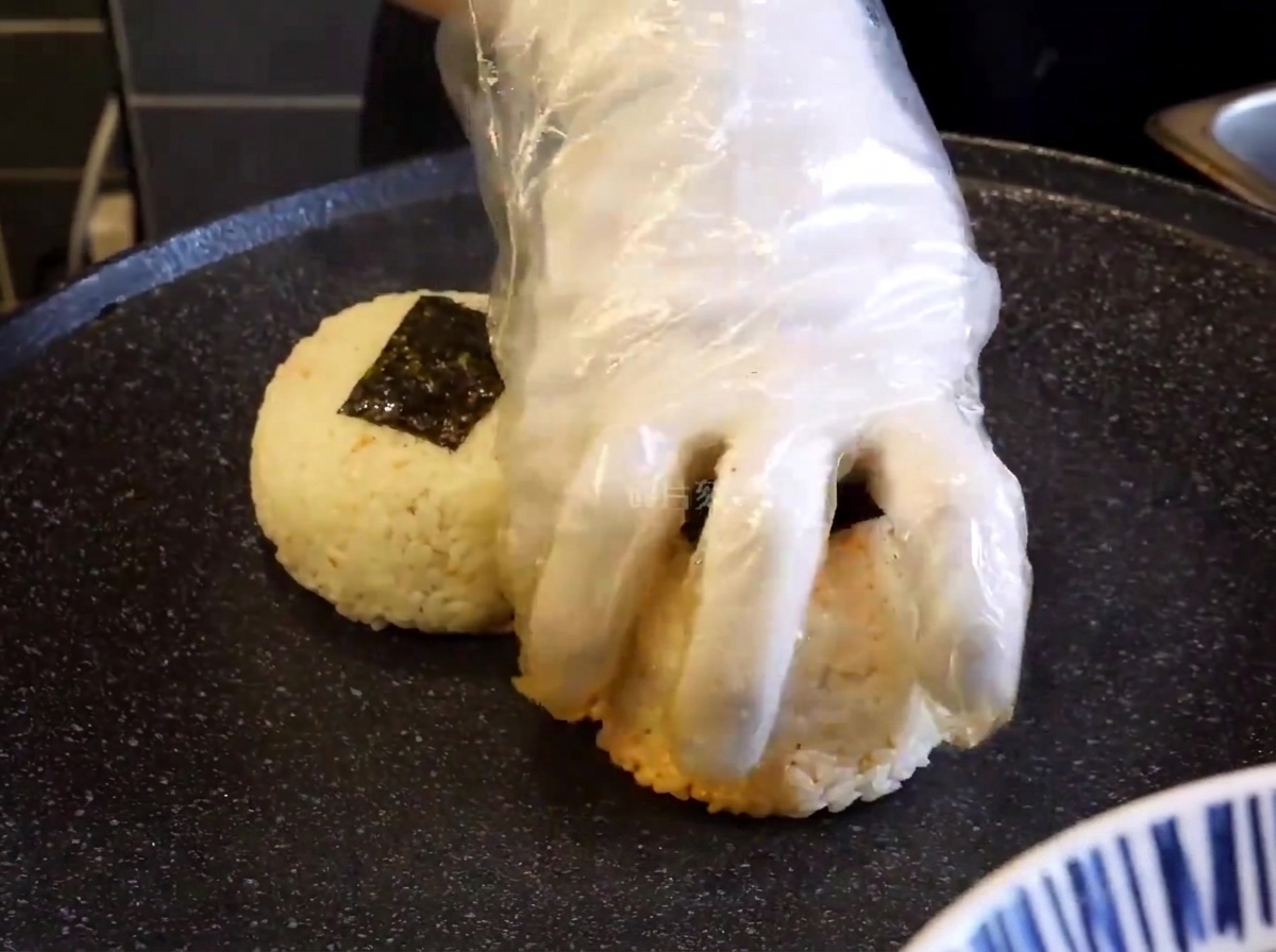 手袋をした手で、グリルの上のおにぎりを押し付けて焼き色をつけている様子。