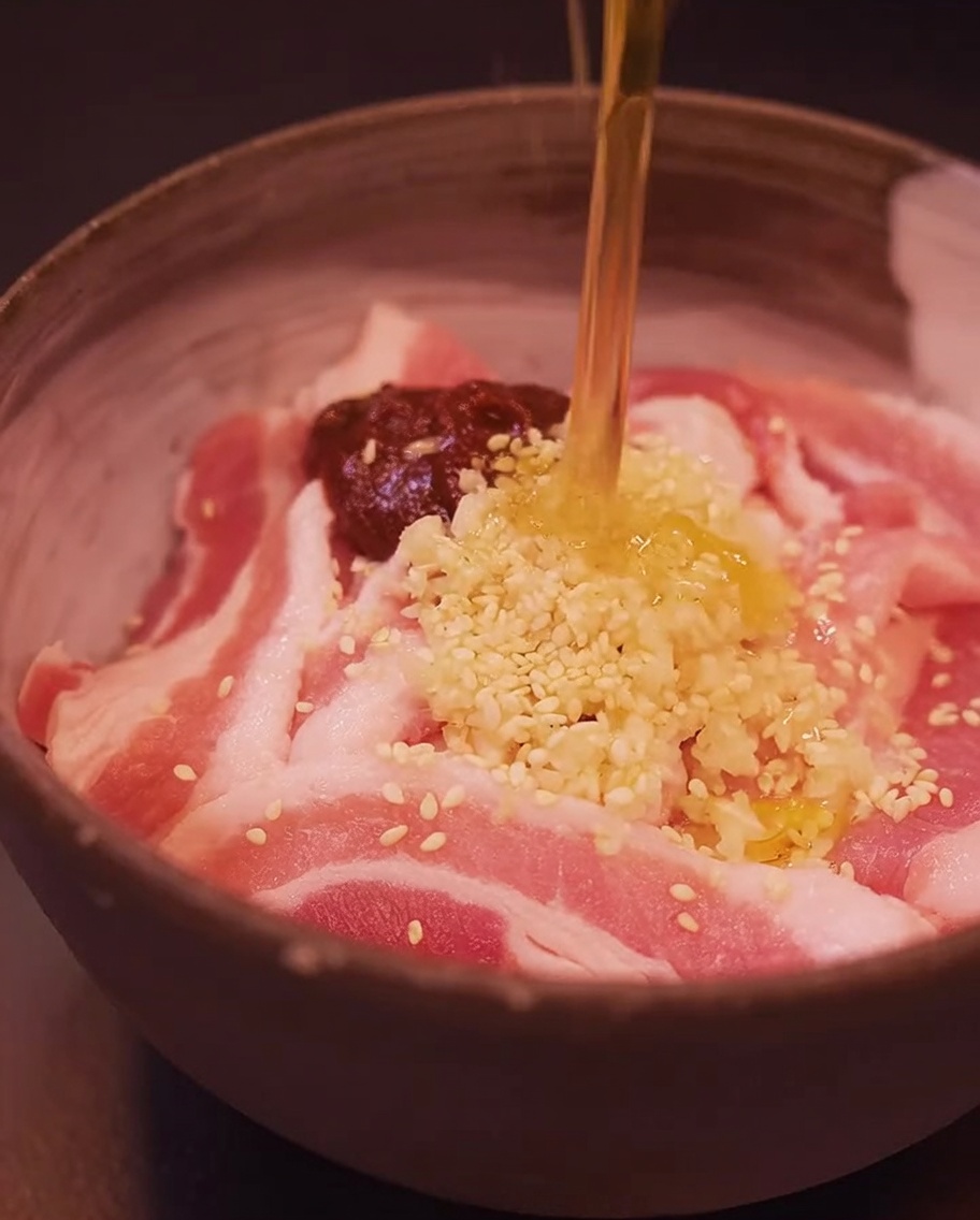 ボウルに入った豚バラ肉に、コチュジャン、おろしにんにく、ごま、ごま油を加えている様子。