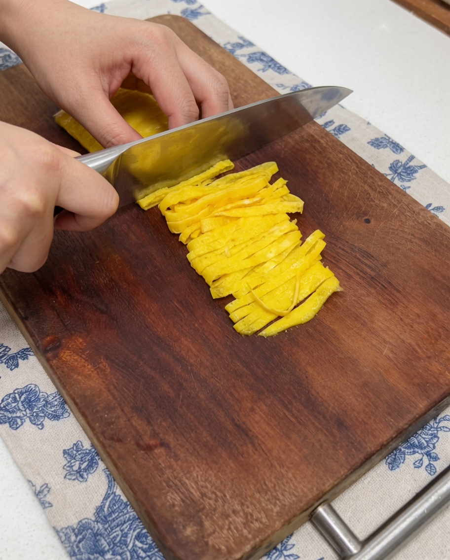 木製のまな板の上で、包丁を使って黄色い薄焼き卵を綺麗に細切りにしている手のクローズアップ。