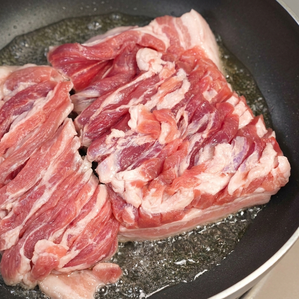 ジュージューと音を立てる豚バラ肉が入ったフライパンと、余分な溶けた脂をスプーンですくい取っている様子。