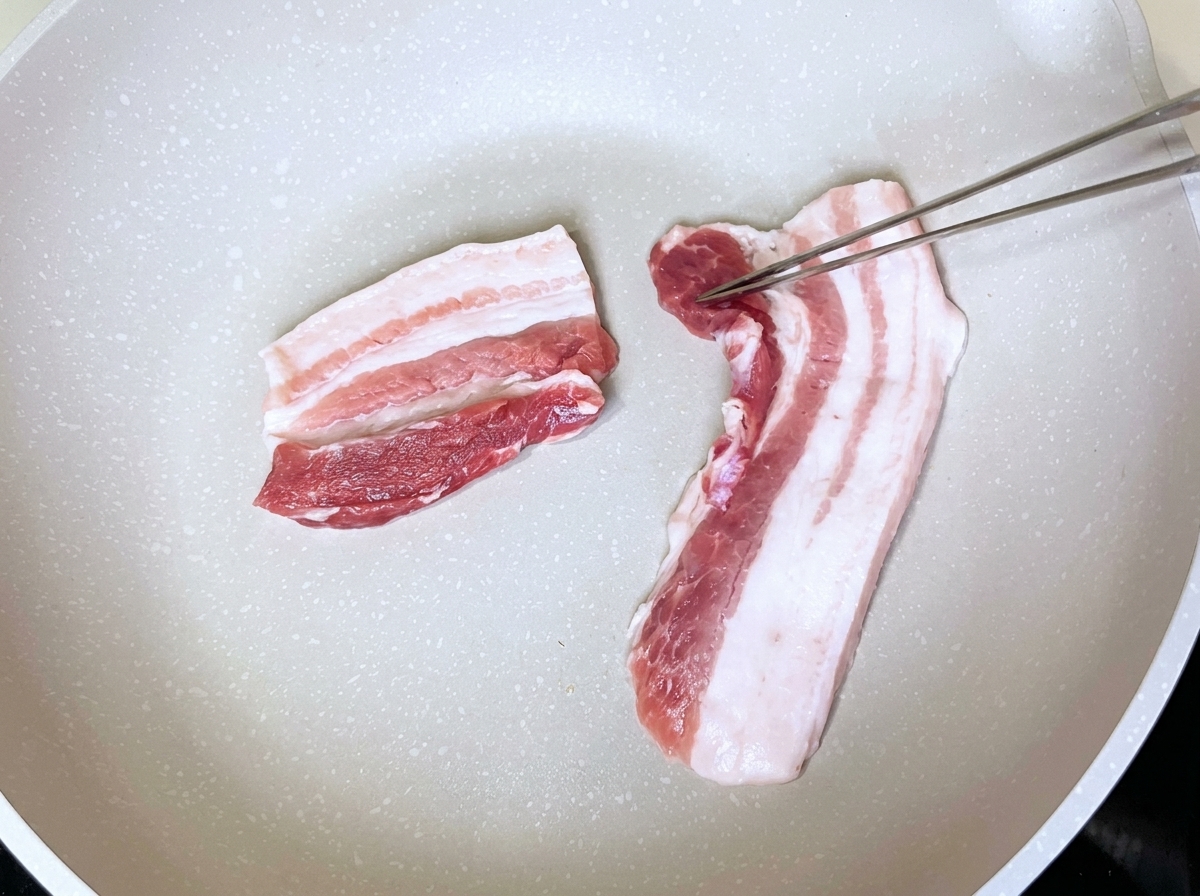 金属製のトングで扱われている、油を引いていない白いフライパンにきれいに並べられた豚バラ肉のスライス。