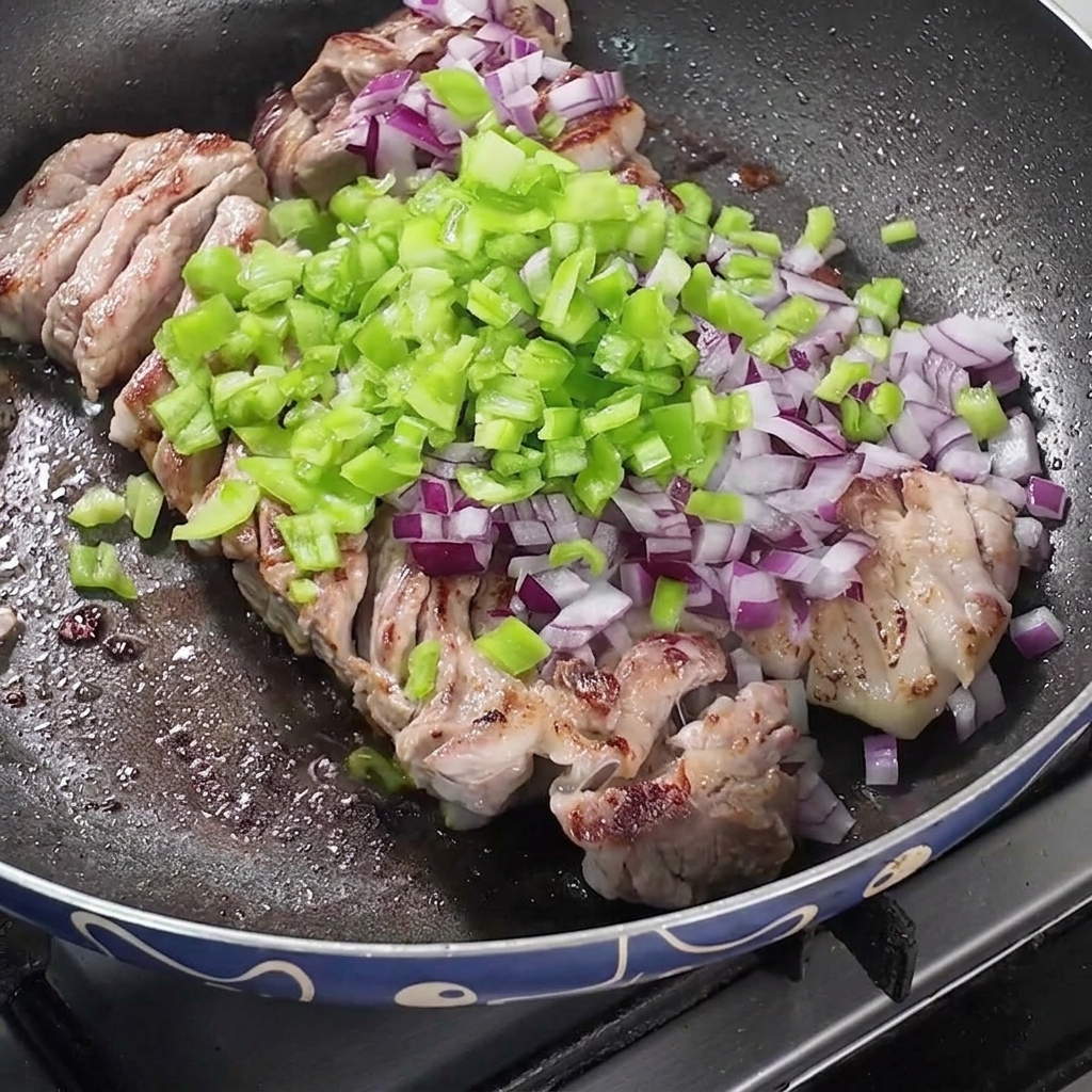 焼いている豚バラ肉の上に、刻んだ赤玉ねぎ、ピーマン、スパイスが乗せられたフライパンの様子。