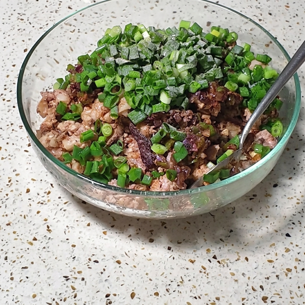 細切り肉と卵の混合物に、新鮮なネギと調味料がトッピングされ、メタルスプーンで混ぜられているガラスボウル。